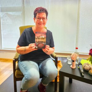 Autorin Evelyn Kühne sitzt auf einem Stuhl, lächelt in die Kamera und hält ihr Buch ‚Für immer an deiner Seite‘ in die Kamera. Neben ihr auf dem Tisch: ein Mikrofon, Notizen und eine Flasche Wasser