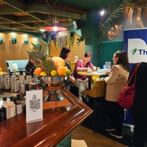 Autorin Evelyn Kühne bei einer Signierstunde in einer stilvollen Bar. Sie sitzt an einem hohen Tisch, signiert Bücher und spricht mit Besucherinnen. Im Vordergrund eine Obstschale auf dem Bartresen.