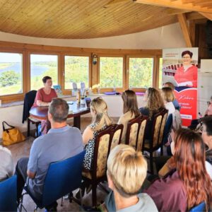 Evelyn Kühne liest vor Publikum in einem lichtdurchfluteten Raum mit Blick auf die Elbe. Die Autorin sitzt an einem runden Tisch, umgeben von Büchern. Neben ihr ein Roll-up mit ihrem Porträt.